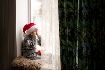 a child in festive clothes and a red Christmas hat sits at the window and covers his face with his hands