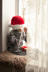 a child in festive clothes and a red Christmas hat sits at the window and covers his face with a hat