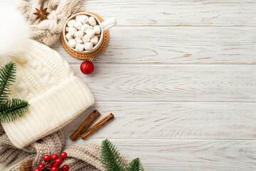 Top view photo of bobble hat red bauble pine branches cup of hot chocolate with marshmallow knitted scarf mistletoe berries anise cinnamon sticks on white wooden table background with copyspace