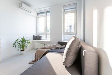 Stylish interior of contemporary bedroom with comfortable furniture. Bed for two with blanket and pillows, mirror and potted plant on gray wall background in modern home with curtains, sun flare