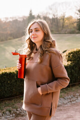Young woman in hoodie holding a red thermo mug in an autumn park. Sunny weather. Fall season.