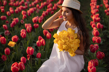 Smiling young lady with bunch of tulips sitting in field at sunset