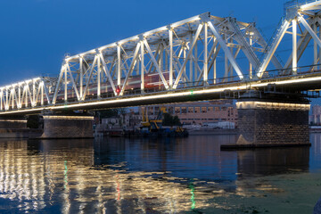Illuminated bridge fragment at night with moving train. Motion blur. Reflection in river. Copy space for text