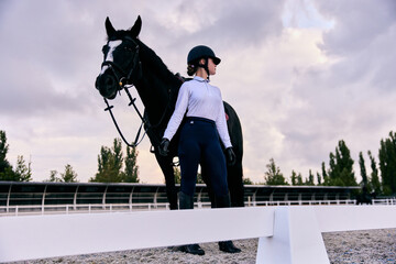 Young sportive girl, professional jockey or horsewoman in sports uniform and helmet with black...