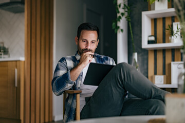 Thoughtful man holding his chin, sitting at home, using a digital tablet, working over it.
