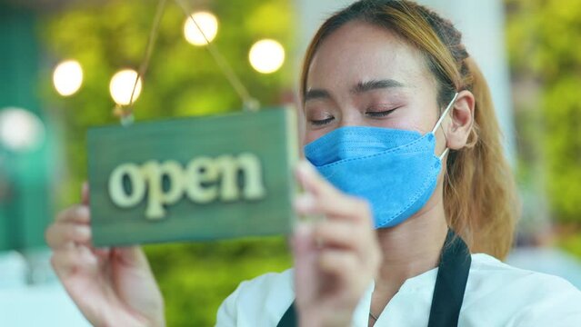 Asian female owner small business wearing protective face mask turning open sign banner.