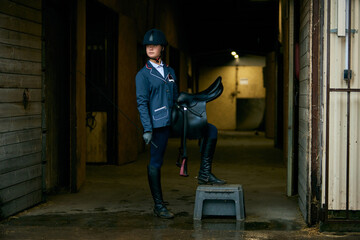 Fototapeta na wymiar Portrait of young girl, jockey and horsewoman in competition uniform and helmet standing stands in stable before the start of horse race