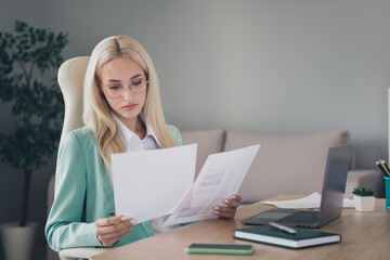 Photo of serious adorable lady wear turquoise jacket working modern device reading papers indoors...