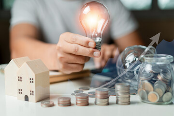 Businessman using calculator to calculate and holding a light bulb and Stacking coins, piggy, wooden house on table. Saving money,accounting, investment, budgeting, and financial planning concept.