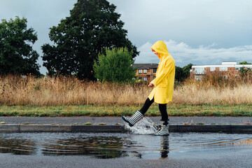 Woman having fun on the street after the rain. Smiling woman wearing rain rubber boots and yellow...