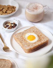 A plate of egg hole toast (toad in a hole) whole bread with snack and a cup of coffee in background, with spoon. A healthy breakfast concept 