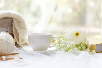 Coffee morning.  White steaming cup of hot coffee for relax after working hobbies crochet on white wooden near window with flowers on autumn and winter season. Lifestyle Concept