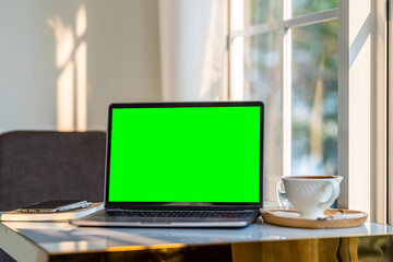 Fototapeta premium Mockup of laptop computer with empty screen with coffee cup and smartphone on table beside the window of the coffee shop background,Green screen