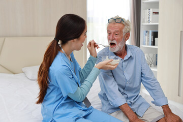 Happy caregiver feeding caucasian senior man on bed with rice porridge soup for breakfast at retirement house. Asian smiling nurse taking good help care and support elder patient at home.