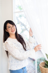 Portrait of Happy Attractive asian people cute woman wearing white lace shirt with the curtains on the window in the room felt like relaxing in house like the background