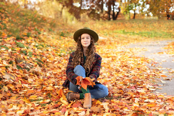 Obraz na płótnie Canvas Young girl in hat and autumn clothes sit on golden fall leaves. Happy girl in green hat and plaid jacket on an autumn background. A smiling girl walking in fall park. 