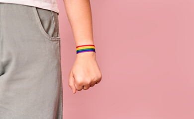 a young woman wearing a bracelet with the colors of the LGBT flag on her arm, her hand clenched into a fist with an LGBT bracelet on it, highlighted on a pink background.