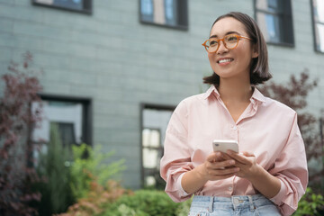Beautiful smiling asian woman wearing stylish eyeglasses holding mobile phone text messaging looking away on the street, copy space. Happy hipster female shopping. Mobile banking concept   