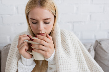 blonde woman in knitted sweater sitting covered in blanket with cup of tea in living room.