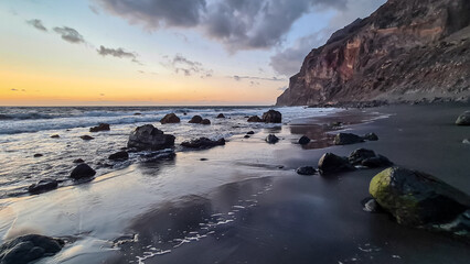 Scenic view during sunset on volcanic black sand beach Playa del Ingles in Valle Gran Rey, La Gomera, Canary Islands, Spain, Europe. Massive cliffs of the La Mercia range. Smooth waves sweeping rocks