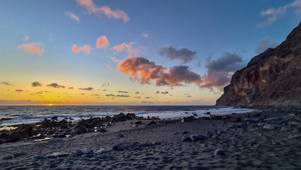 Obraz premium Scenic view during sunset on the volcanic sand beach Playa del Ingles in Valle Gran Rey, La Gomera, Canary Islands, Spain, Europe. Massive cliffs of the La Mercia range. Calm atmosphere at the seaside