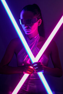Futuristic young woman in trendy mirrored sunglasses with neon sticks in studio