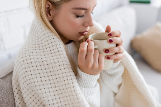 young blonde woman in white sweater sitting covered in blanket with cup of tea.