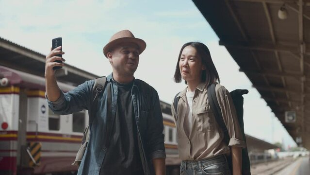 Happy asian handsome man and beautiful woman backpacker wearing hat and backpack walking together in the train station and selfie or facetime for social network with cell phone. 4k resolution.