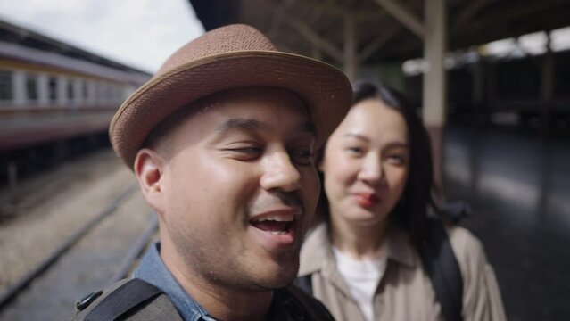 Happy asian handsome man and beautiful woman backpacker wearing hat and backpack walking together in the train station and selfie or facetime for social network with cell phone. 4k resolution.
