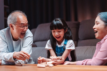 Fototapeta premium Asian happy family of little daughter play toy wooden block in home. Grandparents and young woman loving couple parent having fun play stack tower wood block game at night in living room.