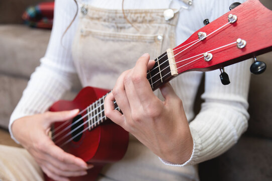 Young woman playing red ukulele at home, sitting on the floor, dressed as a hipster, learning songs