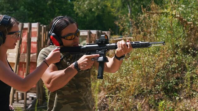 Woman standing next to man aiming submachine gun both wearing safety headphones and goggles. Firearms training at firing range. Outdoor horizontal shot. High quality 4k footage