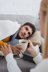 caring girlfriend holding cup of tea near sick boyfriend lying on sofa under blanket.