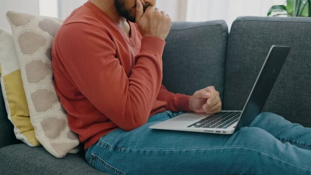 Man frustrated on sofa thinking with laptop, writing online test or assignment for university. Young latino male brainstorming idea in living room typing email, social media or blog post on computer