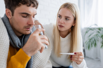 sick man drinking water near worried woman holding digital thermometer.