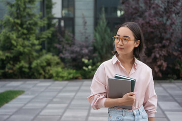 Portrait of smart asian student wearing eyeglasses holding book, laptop standing in university campus, copy space. Education concept 