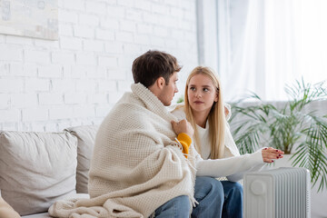 young couple covered in blanket sitting on couch and warming near radiator heater at home.