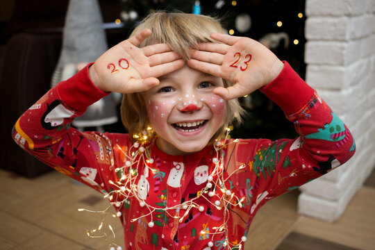 smiling cheerful child in a festive make-up of a deer, numbers of new year 2023 are written on palms. The kid is waiting for a miracle on Christmas Eve. New Year's winter holidays, pampering at home
