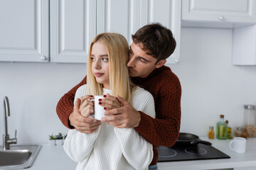 young man in red sweater hugging blonde woman with cup of tea during winter.