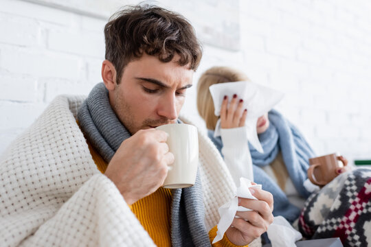 sick man drinking tea and holding tissue near blonde girlfriend in living room. - Powered by Adobe