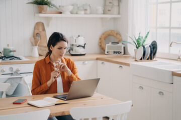 Female housewife working at laptop, makes to-do list, learning language online in kitchen at home