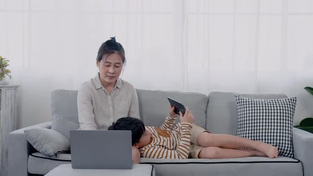 4K, Asian boy playing tablet, He lies on lap Asian single mother, who sitting on sofa working on laptop computer, boy having fun playing games, boy lay in position was most comfortable playing with.
