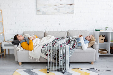 young man and blonde woman holding cups and lying under blankets on sofa near radiator heater.