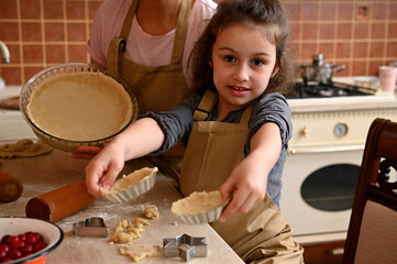 Beautiful little girl and her mother in beige aprons, showing rolled dough in the molds, while making homemade tartlets and tarte. Family fun and cooking concept. Mom and daughter cooking together