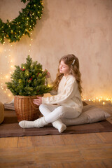 Little cute girl with blond long hair in a warm knitted sweater by a Christmas tree and a big Christmas wreath and burning garlands. Christmas mood 