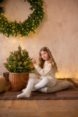 Little cute girl with blond long hair in a warm knitted sweater by a Christmas tree and a big Christmas wreath and burning garlands. Christmas mood 