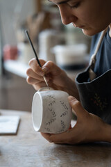 Hands of professional artisan hold white ceramic mug drawing pattern with dark paint using brush on blurred background. Woman enjoys working with handmade pottery craft in studio extreme closeup