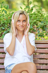 Young woman looking at camera and smiling while enjoying listening music with headphones sitting on a bench in a park. Technology and leisure concept.