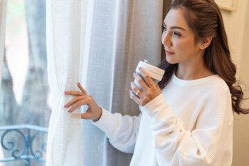 Beautiful Asian woman wakes up early and drinks coffee to refresh her awakening standing by the bedroom window with curtains at home.