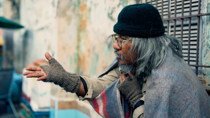  Close-up of a long-haired, dressed Asian man sitting on a wall in an alley, waiting for money and food from passers-by waiting for help. Homeless people who do not have home to sleep on streets - obrazy, fototapety, plakaty